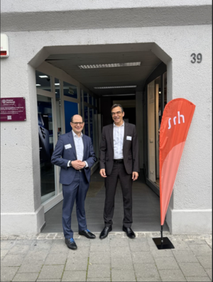 Zwei Männer stehen vor einem Gebäude, neben einem Schild mit der Aufschrift "SRH"